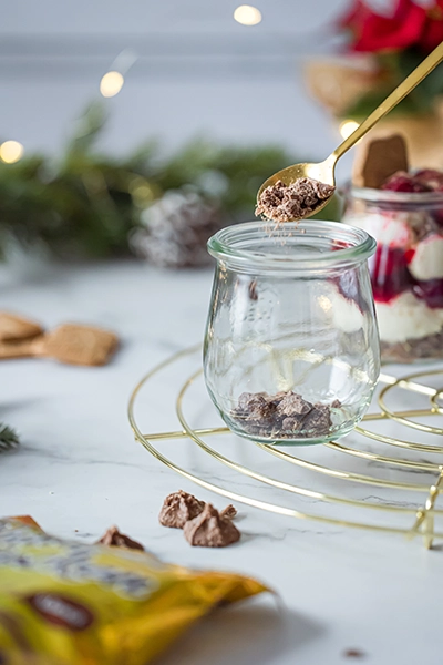 Ein Glas mit Knusperflocken am Boden. Mit einem Löffel wird eine dunkle Creme darauf gefüllt