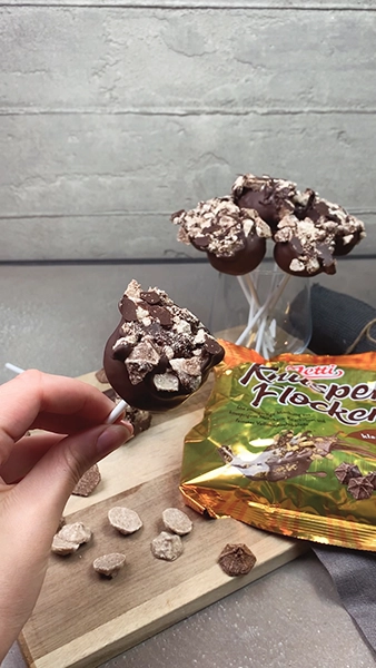 Cakepops mit dunkler Schokolade überzogen und Knusperflocken darauf, stehen auf einem Holzbrett 