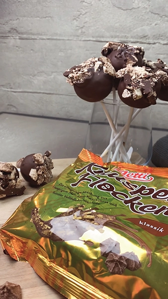 Cakepops mit dunkler Schokolade überzogen und Knusperflocken darauf, stehen auf einem Holzbrett 